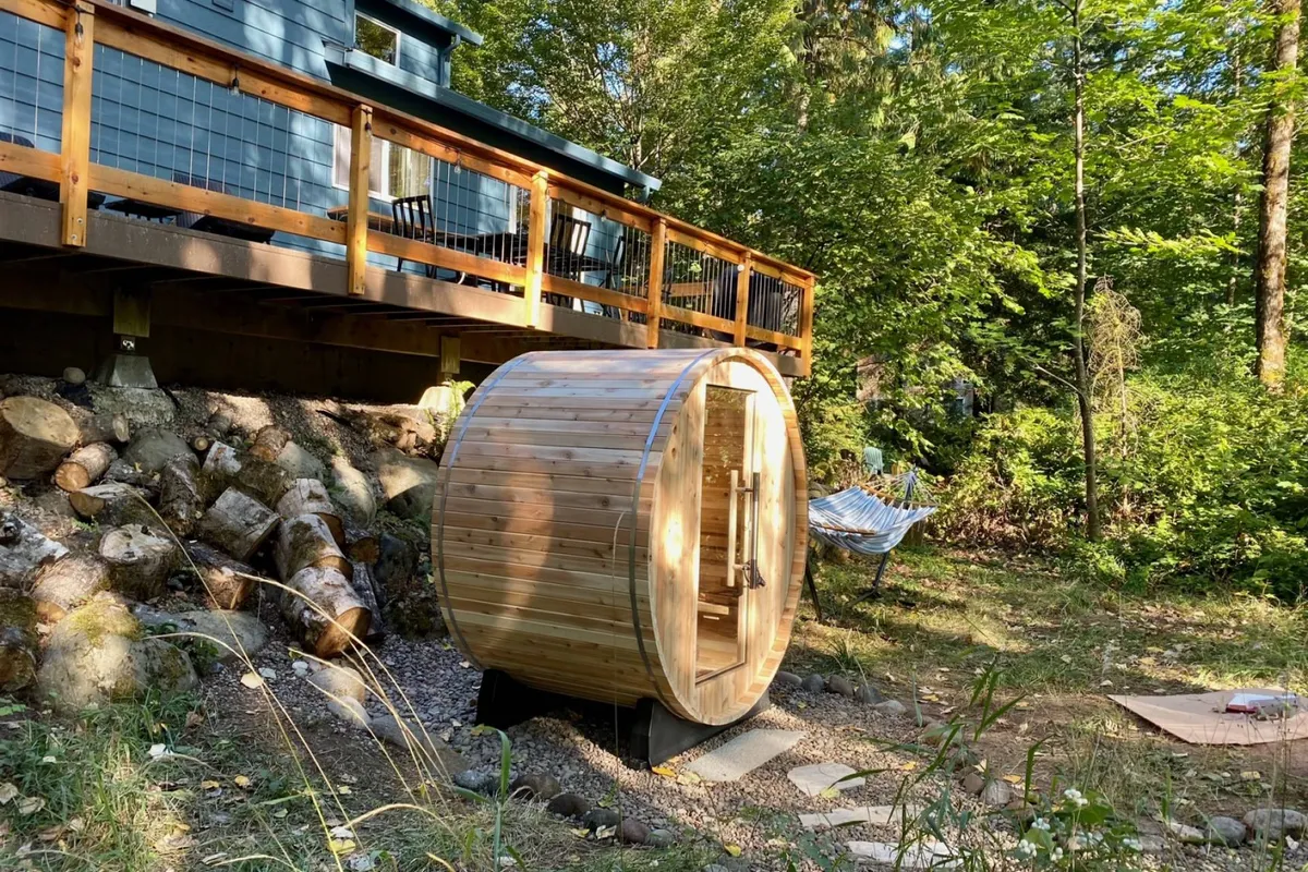 This image features a wooden sauna set on a deck, with a hammock and lush natural surroundings, evoking a restorative, resort-style retreat. The scene is tranquil, with ample space for relaxation and enjoying the serene atmosphere after a day of activities.