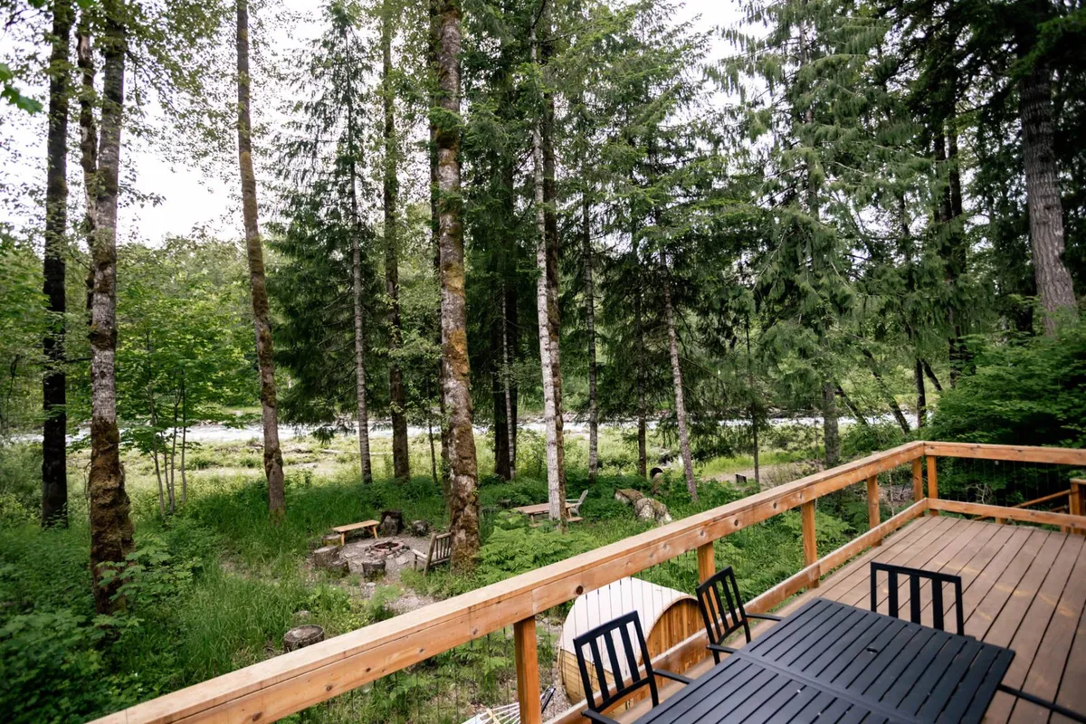 A wide wooden deck set among tall trees, with comfortable seating by a fire pit and the river visible in the background. The area is perfect for outdoor dining, watching sunsets, or listening to the river’s soundtrack, reinforcing the invitation to spill out from the interior for alfresco experiences.