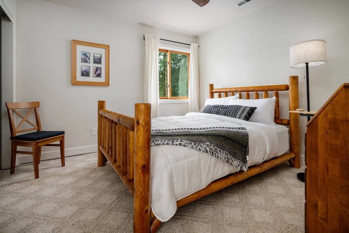A cozy upstairs bedroom with a custom log bed, a soft throw blanket, a lamp for nighttime reading, and a window offering views of lush greenery. The natural materials and abundant light create a restful, inviting setting.