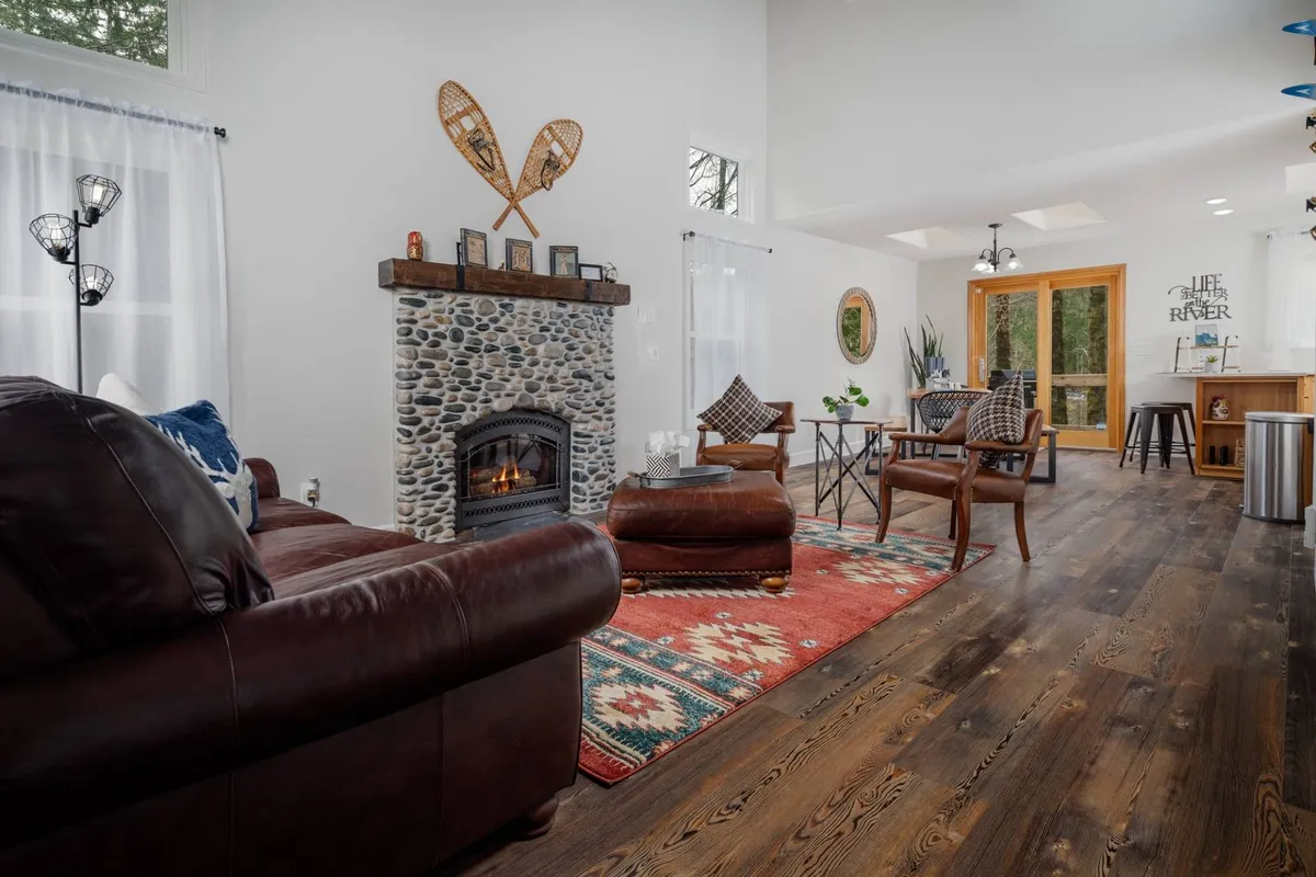 A cozy interior scene featuring a stone fireplace, a plush rug, warm wooden furniture, and large windows that let in plenty of light. The space is set up for both relaxation and socializing, ideal for game nights or cozy mornings with coffee.