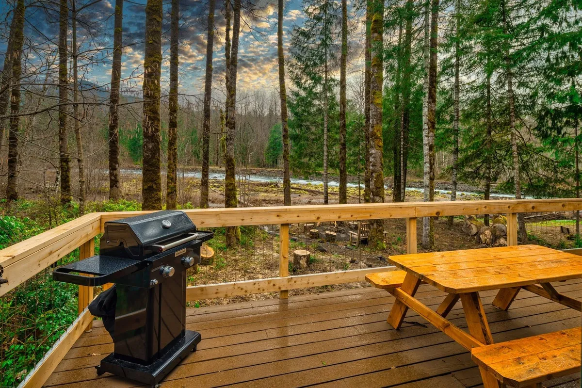 A scenic view of a picnic table and grill set against the backdrop of river views and towering trees. The atmosphere is relaxed, ideal for outdoor dining, gatherings, and enjoying the peaceful environment that defines the resort-style experience.