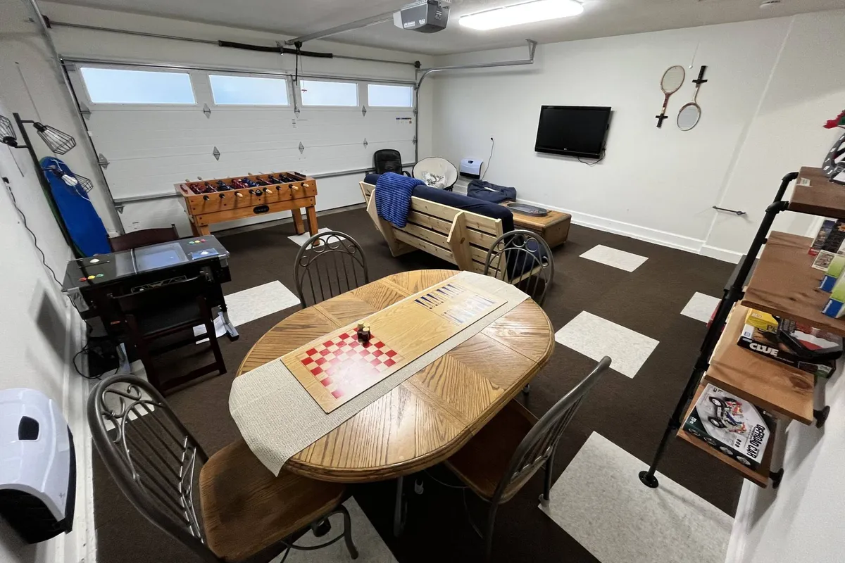 A bright, welcoming game room featuring a round dining table with a checkers board, a comfortable sofa with a blue throw blanket, a foosball table, and a wall-mounted television. The space is set up for various activities, from playing games to lounging and watching TV.