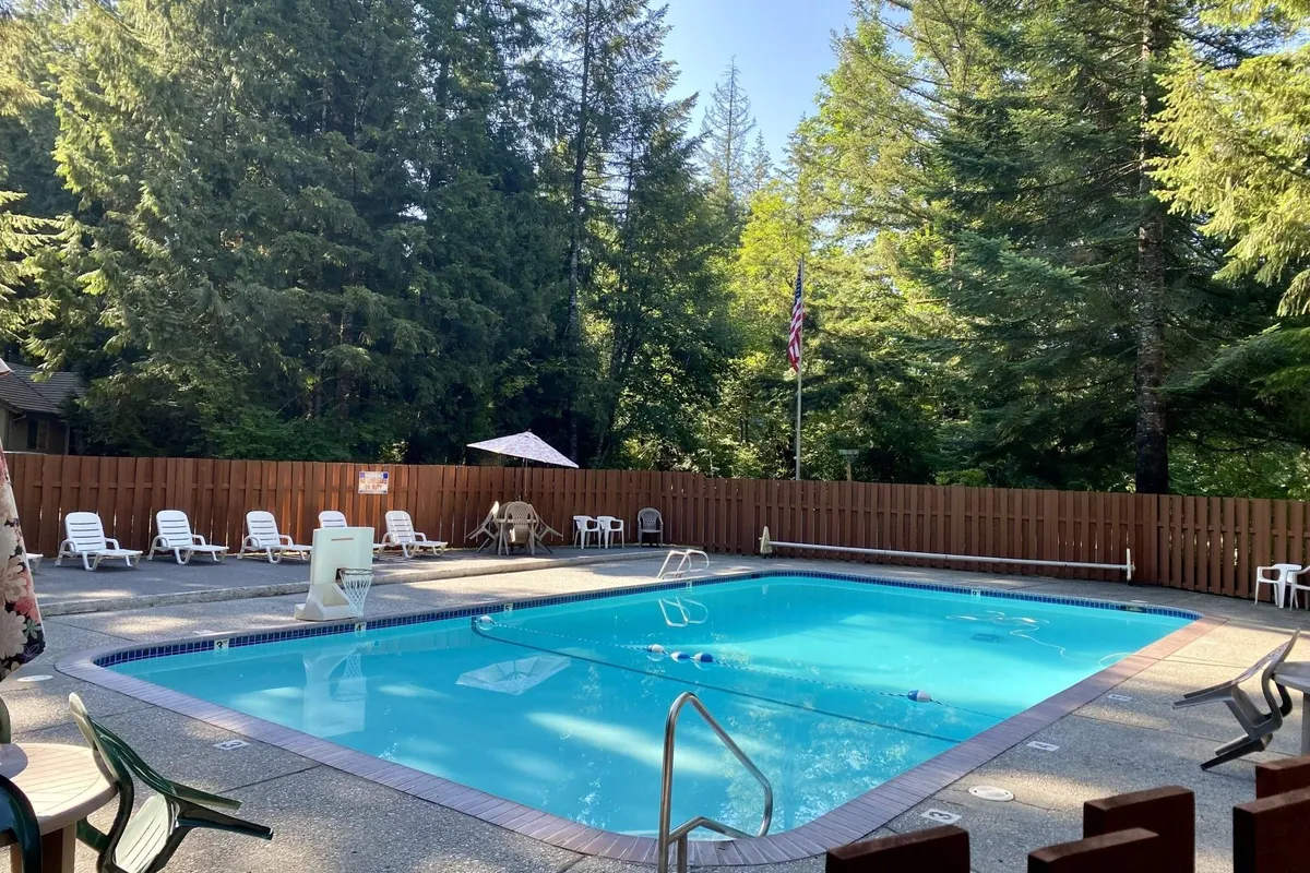 A sparkling swimming pool surrounded by lush trees, with lounge chairs and umbrellas arranged for relaxation. The setting is serene and inviting, suggesting summer leisure and community enjoyment.