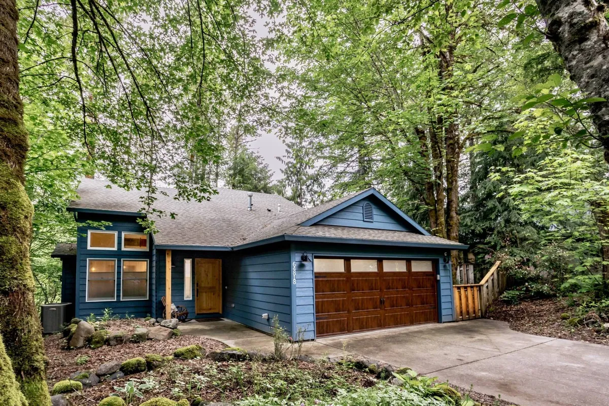 This image displays the blue exterior of the house with a prominent wooden garage door, surrounded by lush greenery. The setting is tranquil and inviting, offering a clear glimpse of the garage's location and its connection to the outdoors.