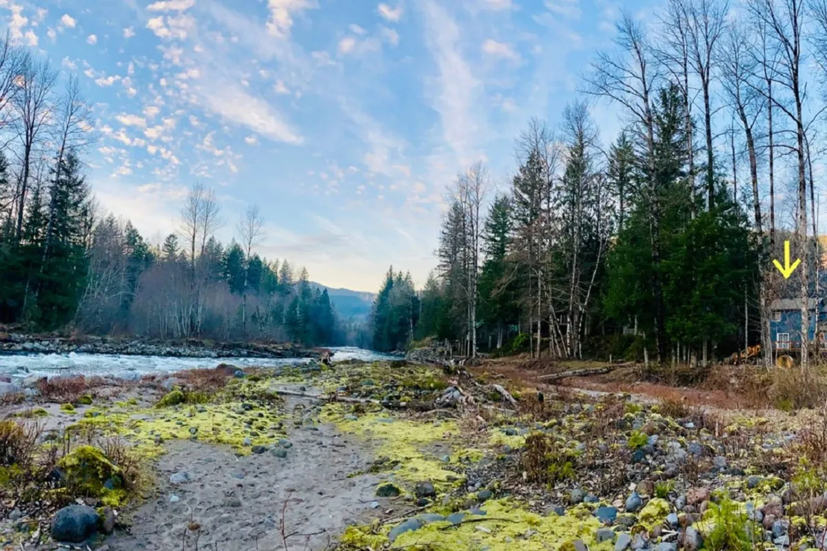 A panoramic view of the river flowing gently along a rocky shore, framed by dense forest and distant mountains. The cabin is visible, nestled within the landscape, emphasizing direct access to the river and natural surroundings.