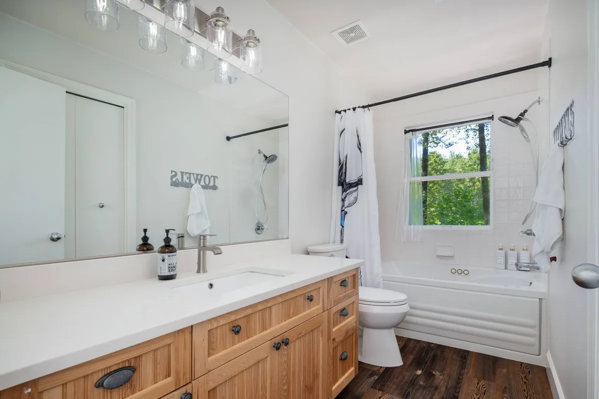 A spa-inspired bathroom featuring a bathtub with a window view, a vanity with wooden cabinetry, a large mirror, and gentle lighting. The space is filled with natural light, enhancing its calm and luxurious atmosphere.