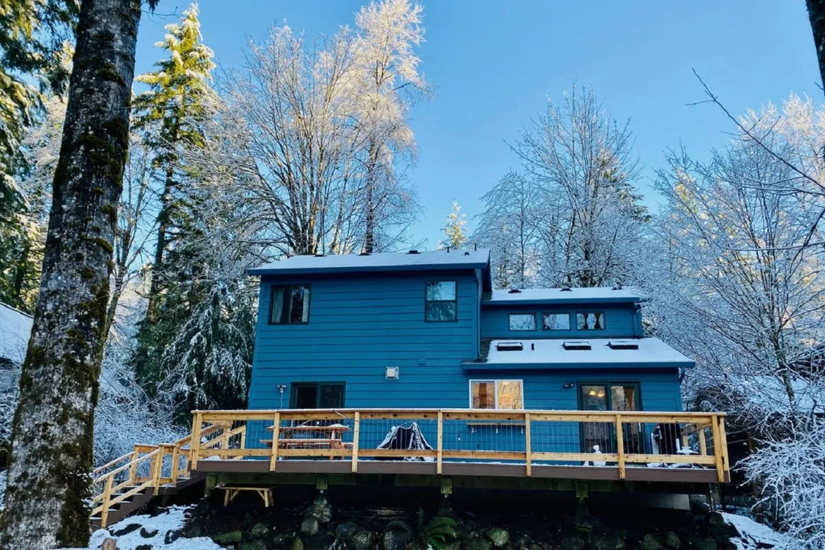 The blue house stands amid snowy trees, with a spacious deck overlooking a wintry landscape. The home’s inviting exterior offers a sense of retreat after mountain sports and outdoor fun.