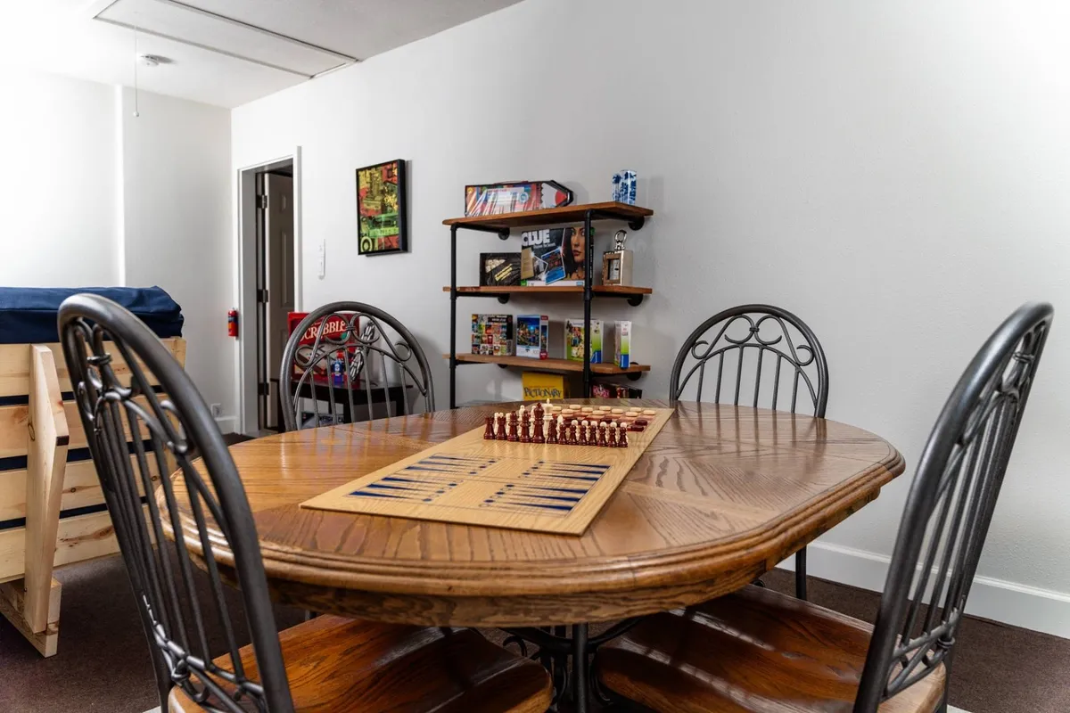 A friendly, well-lit interior focusing on a dining table set up for chess, shelves full of board games, and decorative wall art. The setting radiates warmth and communal energy, clearly geared toward game play and shared activities.