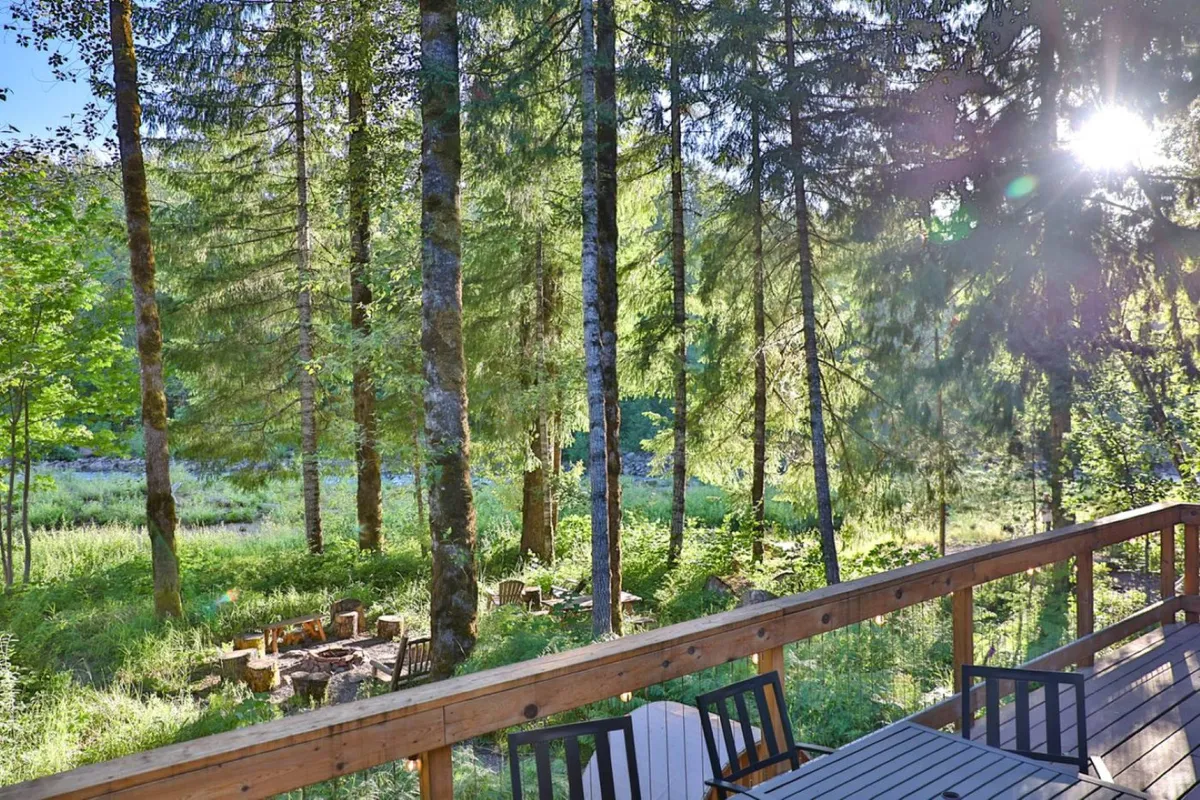A tranquil outdoor space featuring a wooden deck, a custom fire pit surrounded by seating, and tall trees with sunlight filtering through the leaves. The scene evokes a sense of connection to nature and togetherness.