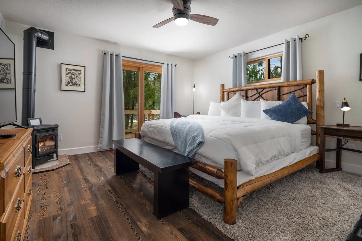 A serene bedroom featuring a large wooden bed dressed in inviting linens, a window that lets in abundant natural light, a rustic wooden dresser, and a cozy fireplace. The soft glow from the fireplace and the view of surrounding greenery create a sense of tranquility, making this space ideal for relaxation and unwinding.