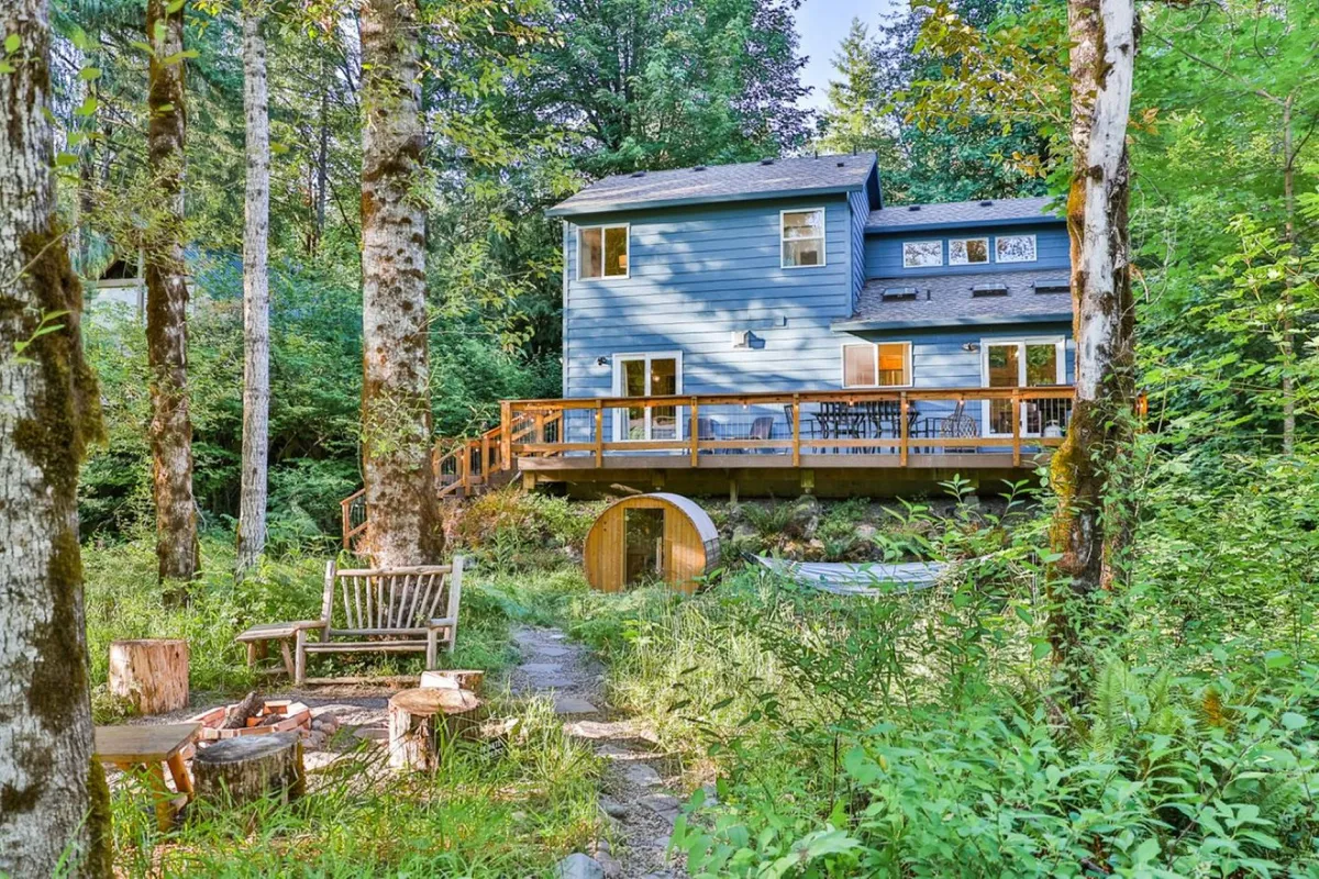 The image showcases a charming blue house nestled among tall trees, set right beside the Sandy River. A spacious wooden deck overlooks the lush surroundings, furnished for outdoor dining and relaxation. The cozy fire pit invites guests to gather and enjoy s'mores or stargazing at night, all bathed in the warm glow of sunset filtering through the forest.