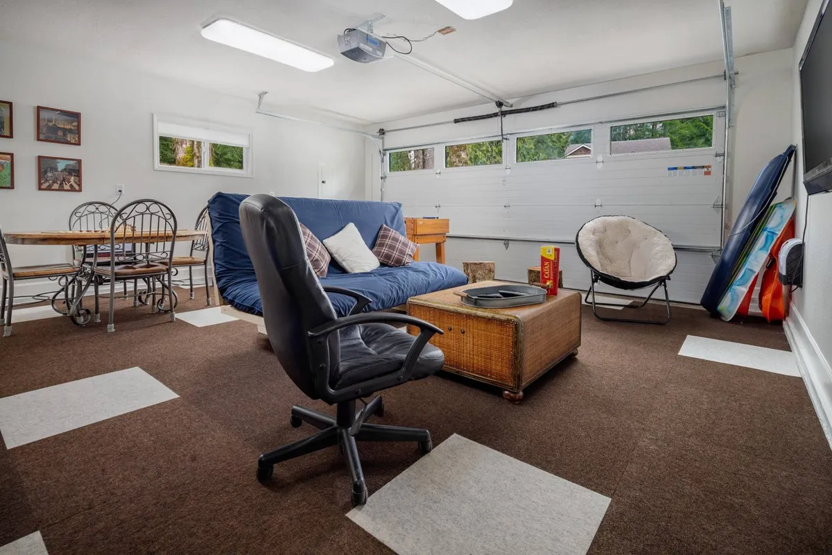 Inside, the converted garage is visible, featuring a cozy sofa, a dining table, an armchair, decorative wall art, and the distinctively styled garage door. This setup suggests a casual, multi-functional space perfect for gatherings, games, and relaxation.