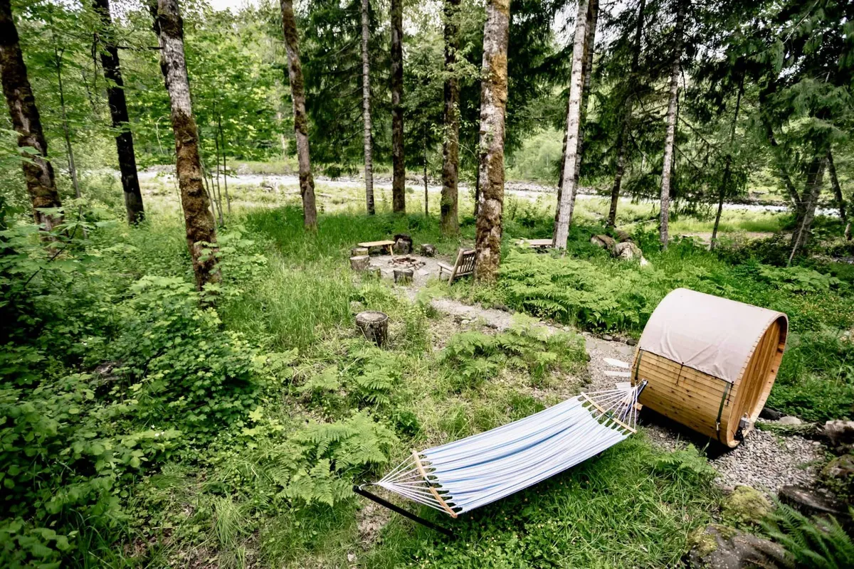 This serene exterior scene features a hammock nestled among dense greenery, a cozy fire pit surrounded by wooden chairs, and a distinctive cedar barrel sauna. The area is inviting and private, ideal for relaxation, wellness rituals, and intimate gatherings.