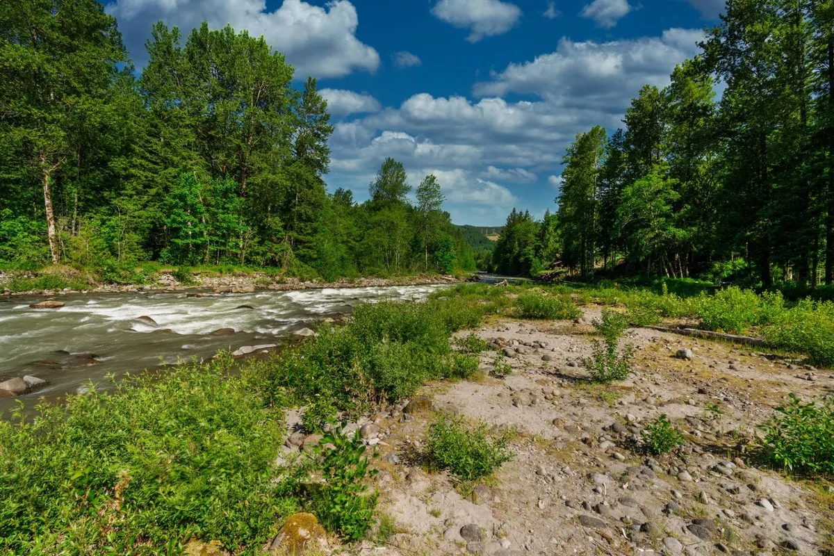 A serene stretch of flowing river bordered by lush greenery and a cloudy sky, with easy access to the water's edge. The tranquil mood invites guests to fish, picnic, or simply relax by the riverside.