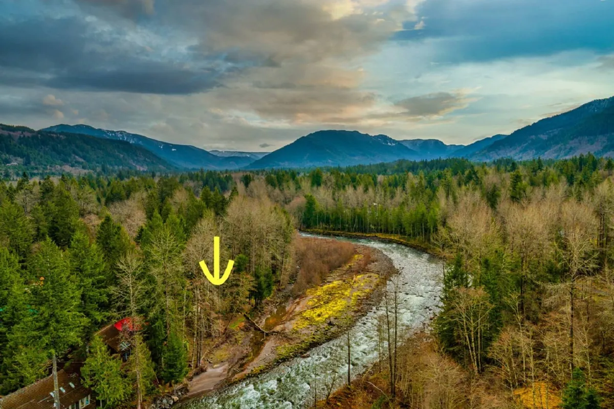 A wide view of a meandering river set against a majestic mountain backdrop and dense forest. The tranquil landscape invites outdoor activities like hiking and fishing, with nature’s beauty on full display as the sun sets over the peaks.